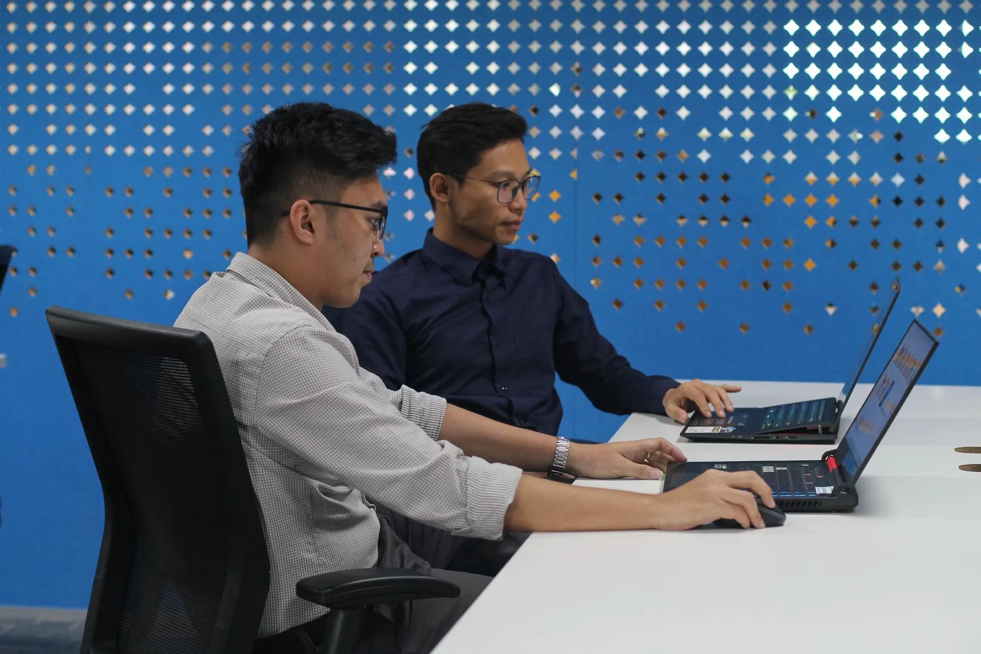 Two designers working on laptops in front of a blue perforated wall